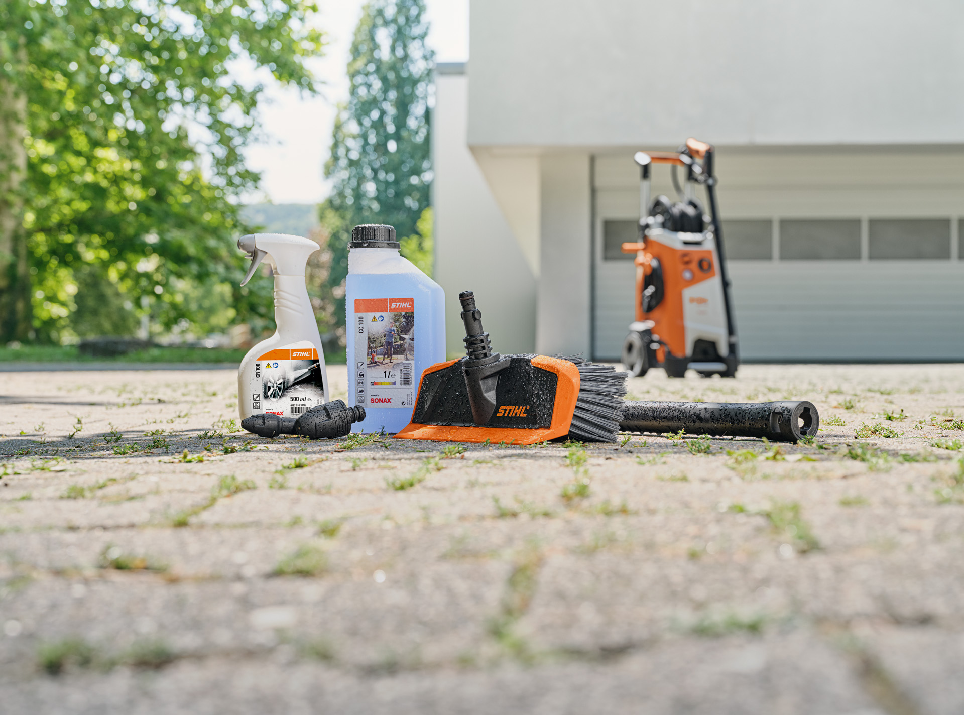  Čistič ráfků kol, autošampon a mycí kartáč STIHL na zemi před bílou garáží. V pozadí vysokotlaký čistič STIHL