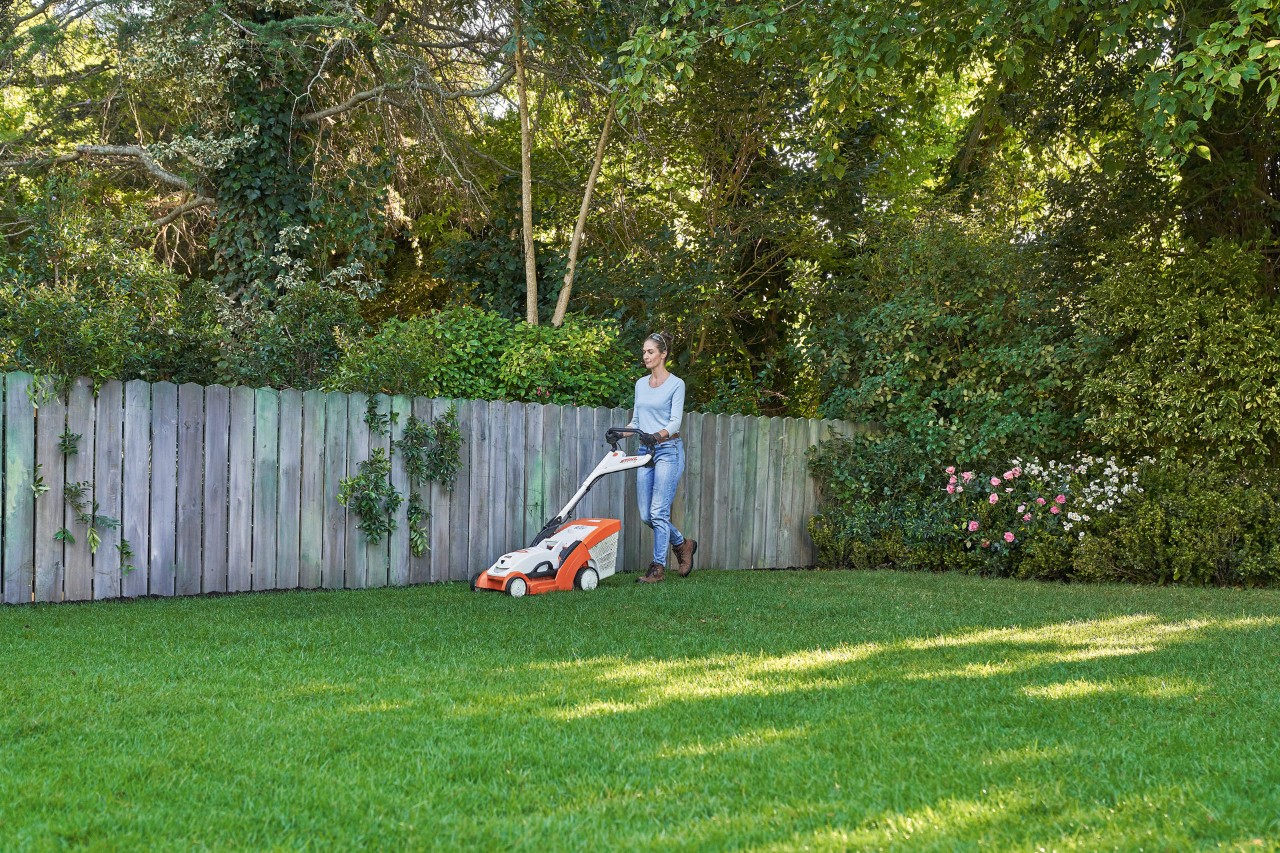 Žena seká zelený trávník před dřevěným plotem pomocí akumulátorové sekačky STIHL RMA 339 C.
