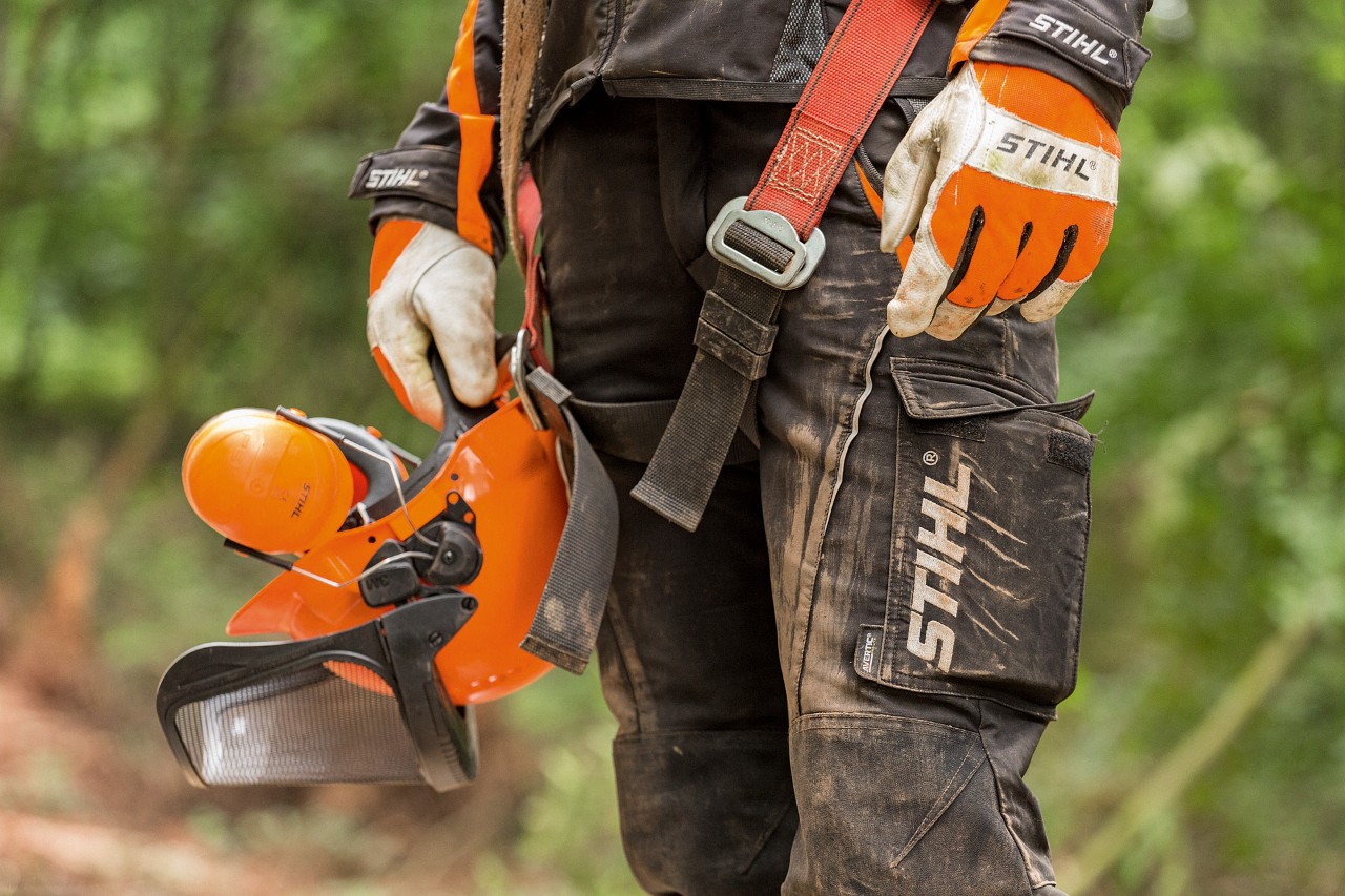 Osoba v kalhotách STIHL s ochranou proti pořezání a s rukavicemi STIHL drží v ruce ochrannou přilbu STIHL 