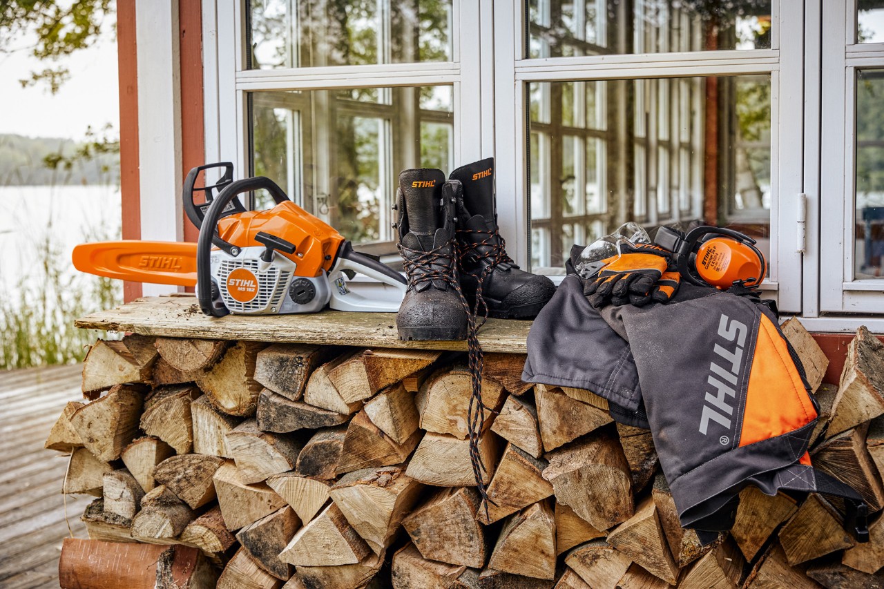 Ochranný oděv STIHL a řetězová pila MS 162 leží na polici nad naskládaným palivovým dřevem