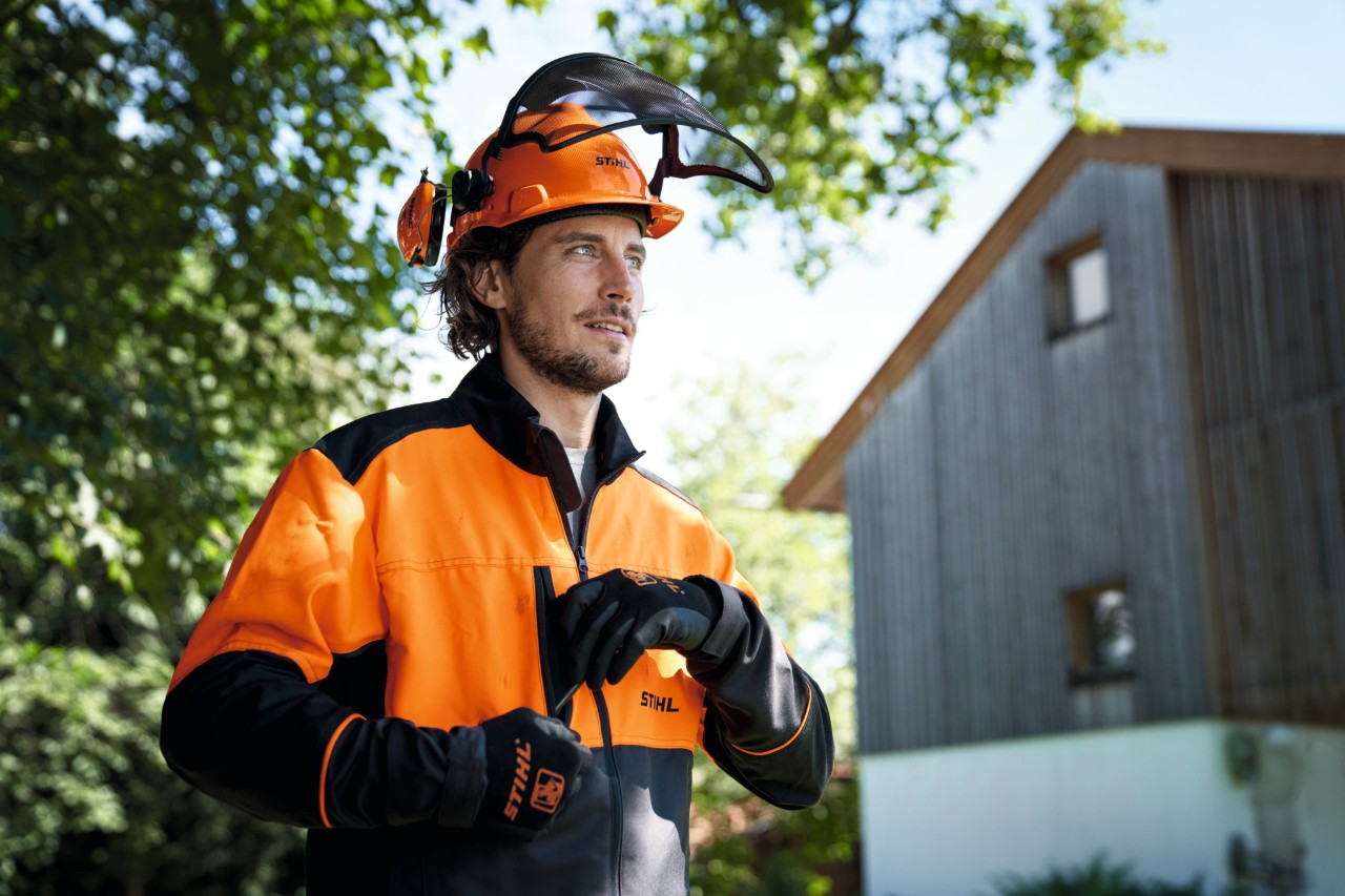 Muž v oranžovo-černém ochranném oděvu STIHL včetně přilby a rukavic se s důvěrou dívá mimo obraz, v pozadí dům a stromy 