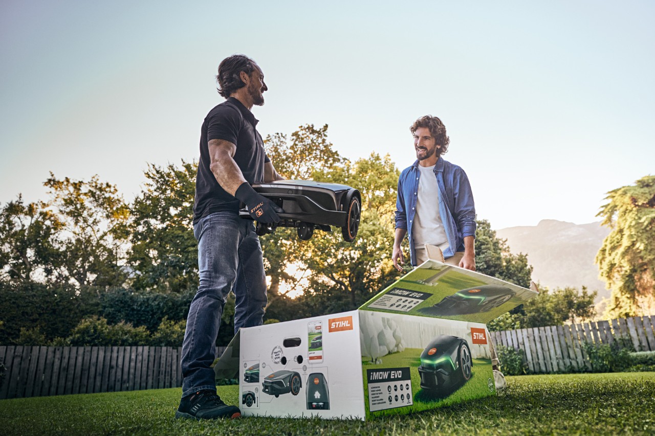 Specializovaný prodejce STIHL stojí na zahradě zákazníka, v ruce drží iMOW® EVO a před sebou má krabici iMOW® a chce pomoci s instalací