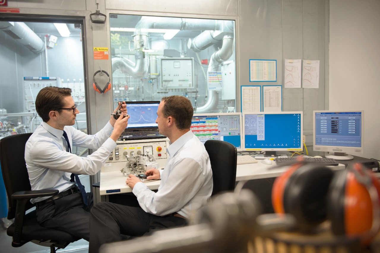 Two men sit in a laboratory at a desk with several monitors and examine part of a new product